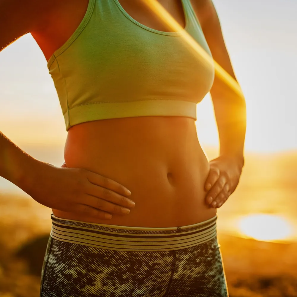 Femme en tenue de sport, mains posées sur le ventre à la lumière dorée du soleil, chakra plexus solaire et écoute de son énergie intérieure