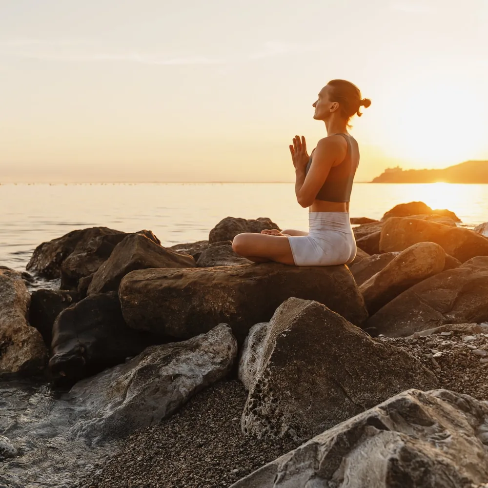 Femme assise en méditation face au soleil levant sur les rochers, chakra plexus solaire et lumière du matin